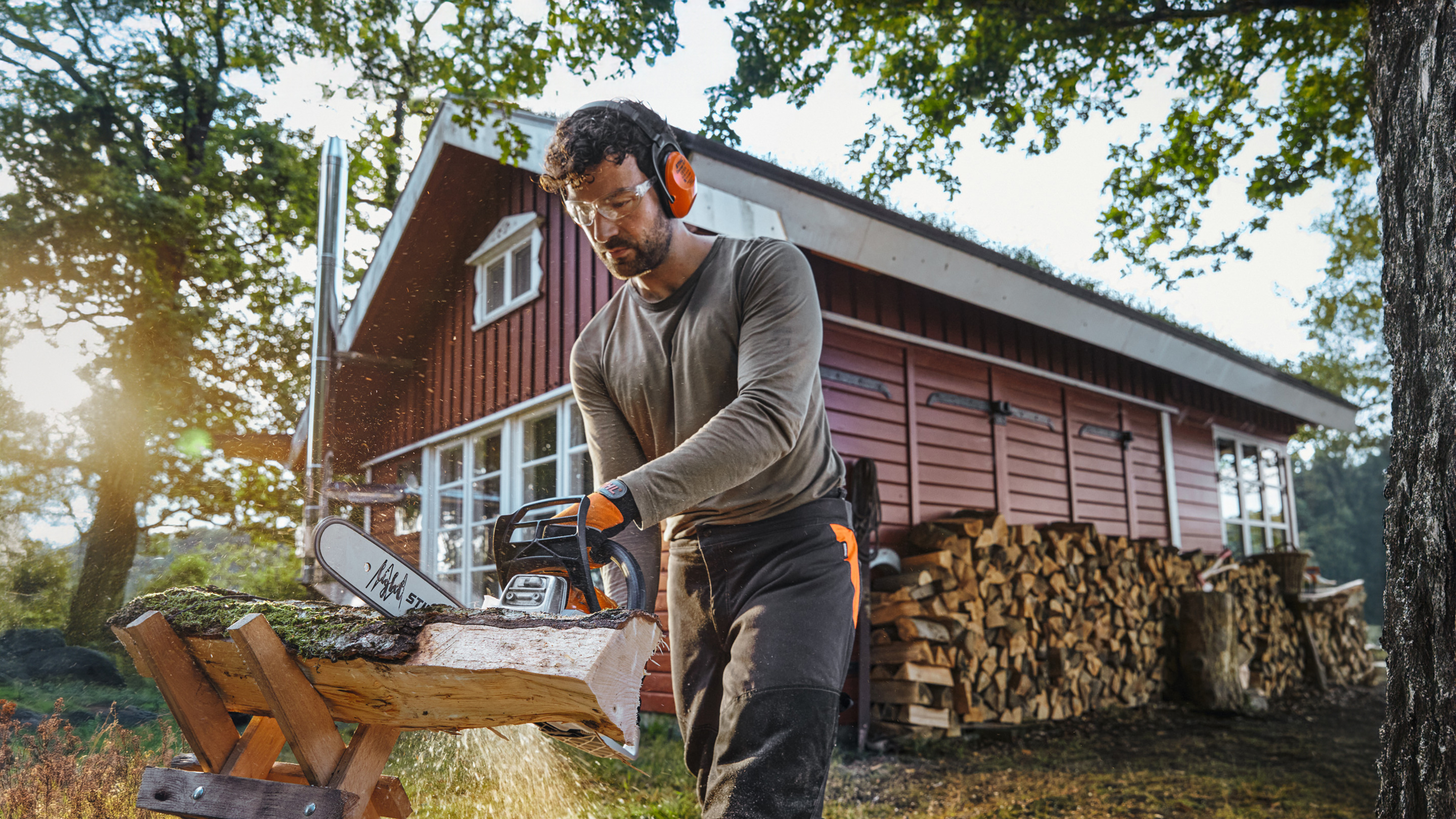 Motosega STIHL con cui un uomo sega un tronco davanti a una capanna di legno rossa con una pila di legna da ardere