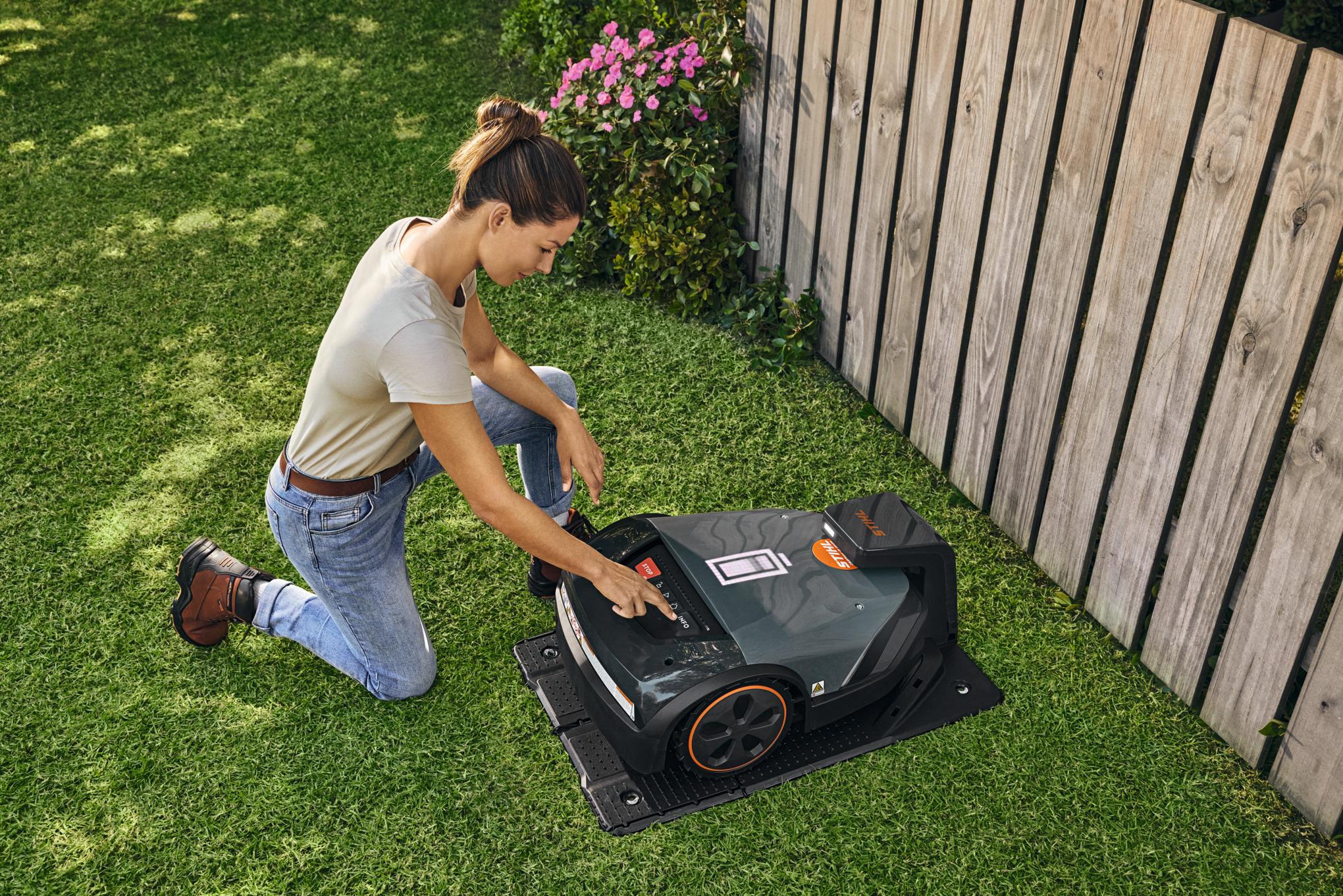 Il tosaerba robotizzato STIHL iMOW® nella sua docking station in giardino mentre una donna regola qualcosa sul tosaerba robotizzato.